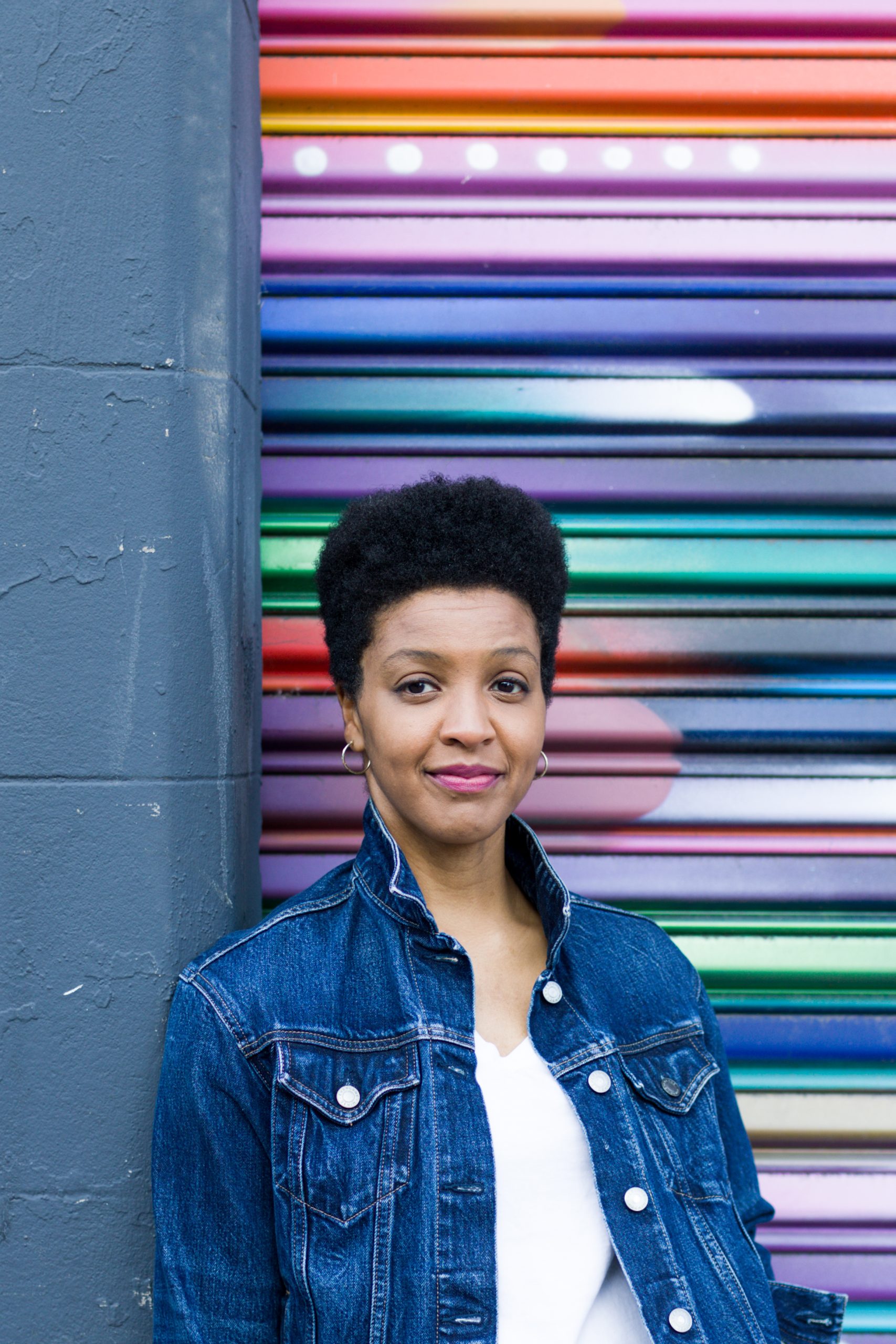 A woman with short black hair in a jean jacket and light shirt in front of a colorful background
