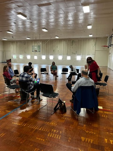 People sitting in a circle on a wooden floor with small square windows in the background.
